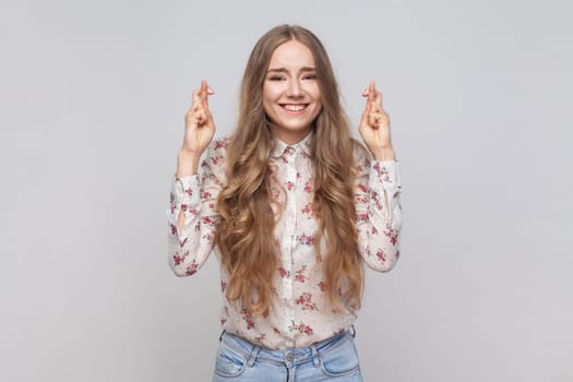 Portrait of hopeful joyous beautiful young adult woman with wavy blond hair raising fingers crossed while making wish, confident to win. Indoor studio shot isolated on gray background.