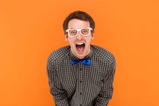 Emotional scared man nerd looks with frightened expression, looks nervously at camera, demonstrates phobia, wears shirt with blue bow tie and glasses. Indoor studio shot isolated on orange background.