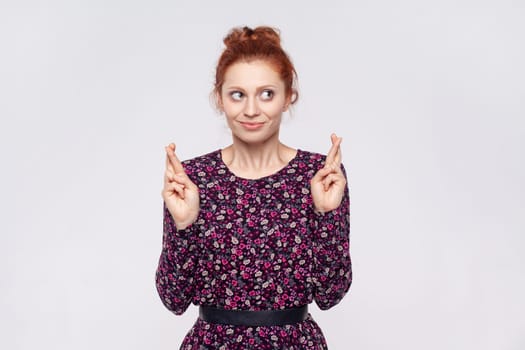 Portrait of redhead woman wearing dress makes wish believes in good luck, hopes dreams come true, has hopeful expression, anticipates for something. Indoor studio shot isolated on gray background.