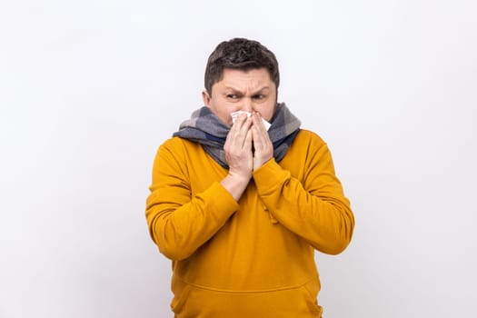 Man in warm scarf coughing sneezing hard in napkin, feeling unwell with runny nose, severe seasonal allergy, flu symptoms, wearing urban style hoodie. Indoor studio shot isolated on white background.