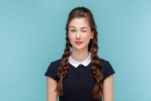 Portrait of coquettish happy positive woman with braids standing and looking winking to camera, flirting with somebody, wearing black dress. woman Indoor studio shot isolated on blue background.