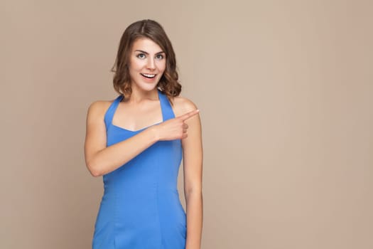Portrait of woman with wavy hair showing something aside at blank wall, being in high spirit, advertisement area, wearing blue dress. Indoor studio shot isolated on light brown background.