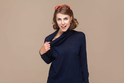 Portrait of beautiful attractive woman with wavy hair in red red glasses on her head standing looking at camera, keeps hand on her collar. Indoor studio shot isolated on light brown background.