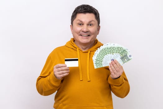 Positive middle aged man holding and showing euro cash and credit card, satisfied with cashback, currency exchange, wearing urban style hoodie. Indoor studio shot isolated on white background.