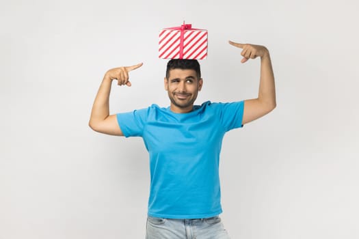 Portrait of optimistic positive unshaven man wearing blue T- shirt standing with gift box on his head, pointing at present, having funny expression. Indoor studio shot isolated on gray background.