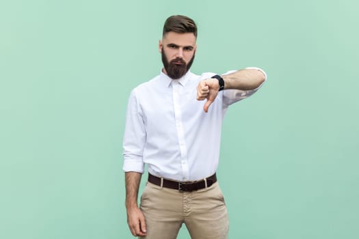 Bearded businessman wearing shirt criticizing bad quality with thumbs down displeased grimace, showing dislike gesture, expressing disapproval. Indoor studio shot isolated on light green background.