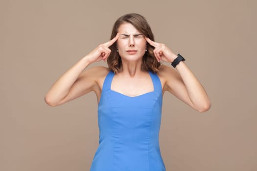 Sick unhealthy woman with wavy hair keeps fingers on temples, suffering terrible headache, standing with closed eyes, wearing blue dress. Indoor studio shot isolated on light brown background.