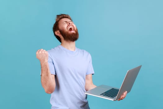Portrait of handsome amazed satisfied positive bearded man holding laptop in hands, clenching fist, looking up with open mouth and triumphing. Indoor studio shot isolated on blue background.