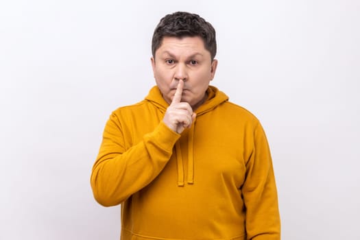 Portrait of middle aged man holding finger on lips making hush silence gesture, asking to keep secret, don't speak, wearing urban style hoodie. Indoor studio shot isolated on white background.