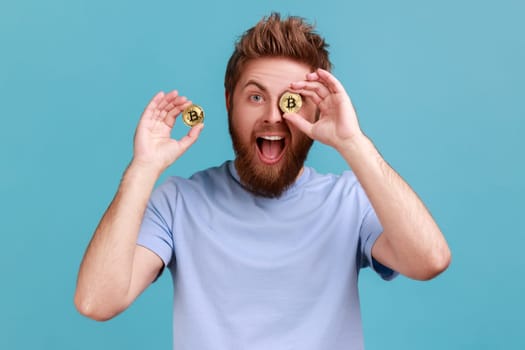 Portrait of excited positive bearded man with open mouth covering eye with golden btc coin and smiling to camera, advertising of cryptocurrency. Indoor studio shot isolated on blue background.