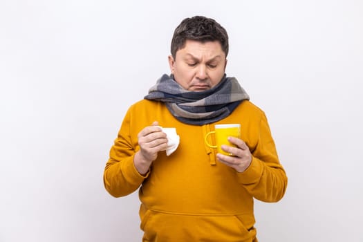Home treatment. Flu-sick man in scarf holding mug and tissue for runny nose, taking medicine with hot tea, suffering fever, wearing urban style hoodie. Indoor studio shot isolated on white background.