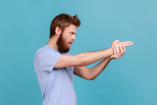 Side view of focused bearded man pointing finger gun gesture to target, threatening to kill, shooting with hand pistol and looking menace. Indoor studio shot isolated on blue background.
