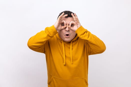 Portrait of curious man looks through fingers in binoculars gesture, observing distant with astonished look, watching afar, wearing urban style hoodie. Indoor studio shot isolated on white background.