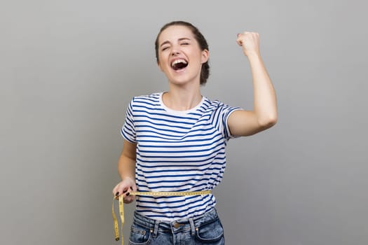 Portrait of extremely happy woman in striped T-shirt holding on waist tape, expressing excitement, rejoicing, clenched fist, screaming with happiness. Indoor studio shot isolated on gray background.