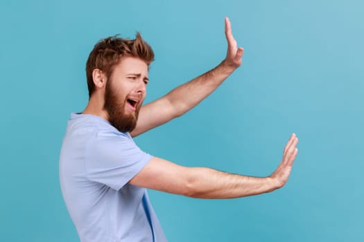 Side view of shocked scared unhappy young bearded man screaming in horror and fear, keeping hands raised to defend himself, panic attack. Indoor studio shot isolated on blue background.