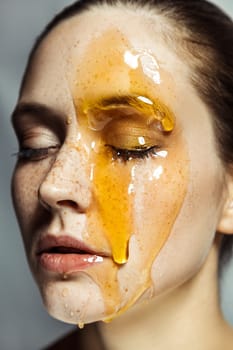 Closeup portrait of beautiful calm young brunette woman with freckles, doing cosmetology procedures, applying honey on her face. Indoor studio shot isolated on gray background.