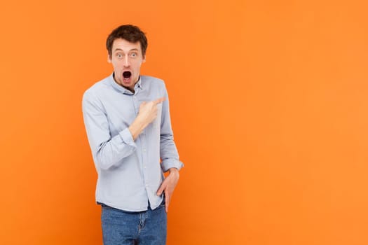 Shocked amazed young adult man pointing aside with finger, showing copy space for advertisement or promotional text, wearing light blue shirt. Indoor studio shot isolated on orange background.