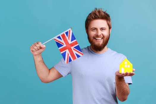 Portrait of happy bearded man standing with paper house and flag of Great Britain, concept of immigration and house purchase in England. Indoor studio shot isolated on blue background.