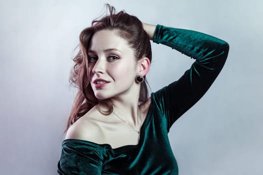 Beauty portrait of smiling cunning attractive woman wearing green dress keeps hand on her hair, looking at camera confident expression. Indoor studio shot isolated on gray background.