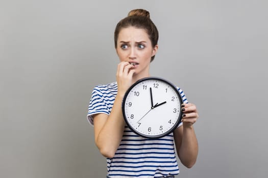 Portrait of woman wearing striped T-shirt holding wall clock in hands, biting nails, looking at camera with nervous expression, deadline. Indoor studio shot isolated on gray background.