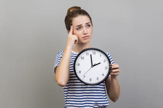 Portrait of pensive thoughtful woman wearing striped T-shirt holding wall clock, thinking about deadline, looking away, keeps finger on head. Indoor studio shot isolated on gray background.