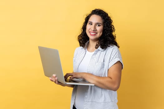 Portrait of satisfied positive happy woman with dark wavy hair working on laptop, looking at camera screen with pleasant expression. Indoor studio shot isolated on yellow background.