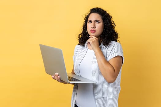 Portrait of pensive thoughtful woman with dark wavy hair working on laptop, looking away and holding chin, thinking about her work. Indoor studio shot isolated on yellow background.