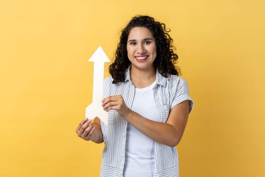 Portrait of smiling satisfied woman with dark wavy hair pointing up looking at camera with smile, growth and increase concept. Indoor studio shot isolated on yellow background.