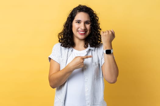 Portrait of positive woman with dark wavy hair standing, looking at camera, pointing at her smart watch, expressing happiness. Indoor studio shot isolated on yellow background.