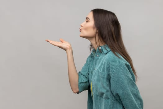 Catch my kiss. Side view of lovely romantic young woman looking ahead air kissing over palms hands, sending love, wearing casual style jacket. Indoor studio shot isolated on gray background.