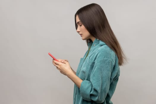 Side view of serious pretty woman reading message on smartphone, using mobile device for communication, browsing web, wearing casual style jacket. Indoor studio shot isolated on gray background.