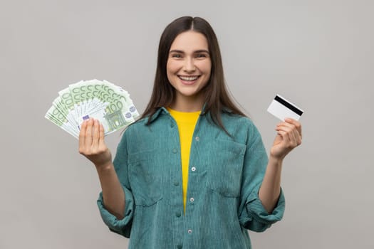 Happy satisfied young woman holding credit card and fan of euro cash looking at camera with toothy smile, wearing casual style jacket. Indoor studio shot isolated on gray background.