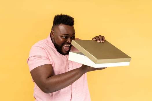 Portrait of optimistic man wearing pink shirt opening gift box and peeping inside with toothy smile, satisfied with present. Indoor studio shot isolated on yellow background.