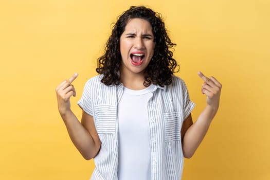 Portrait of woman with dark wavy hair rejecting communication, showing middle fingers to express disrespect and hate, screaming. Indoor studio shot isolated on yellow background.