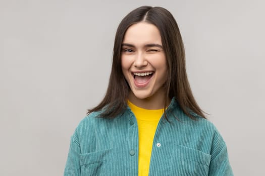 Portrait of good looking cheerful woman winking eye and flirting with someone, looks at camera, expressing happiness, wearing casual style jacket. Indoor studio shot isolated on gray background.
