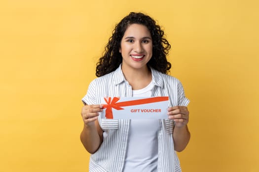 Portrait of delighted happy satisfied woman with dark wavy hair holding gift voucher, holiday gift card, coupon and certificate. Indoor studio shot isolated on yellow background.
