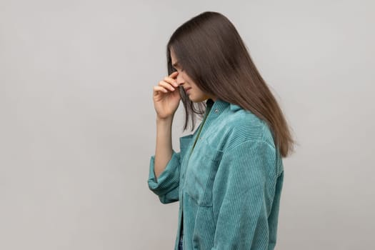 Side view of depressed woman holding her head down, touching face and crying with dramatic grimace, feeling sorrow regret, wearing casual style jacket. Indoor studio shot isolated on gray background.