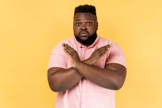 No way, absolutely not. Portrait of amazed man wearing pink shirt showing x sign with crossed hands, meaning stop, this is the end. Indoor studio shot isolated on yellow background.
