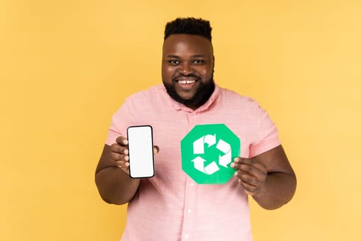 Portrait of man wearing pink shirt showing mobile phone with blank screen and recycling symbol, being happy, looking at camera with toothy smile. Indoor studio shot isolated on yellow background.