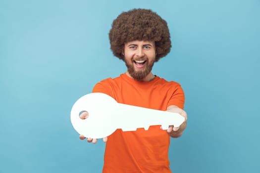 Portrait of man with Afro hairstyle wearing orange T-shirt holding in hand big paper key as symbol of her own apartment, state housing program. Indoor studio shot isolated on blue background.