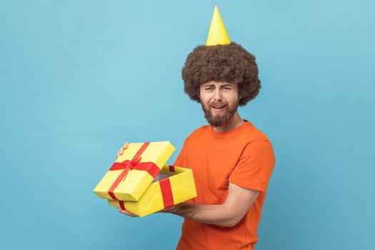 Portrait of sad upset man with Afro hairstyle wearing orange T-shirt and party cone holding present box, opened gift, feeling sorrow. Indoor studio shot isolated on blue background.