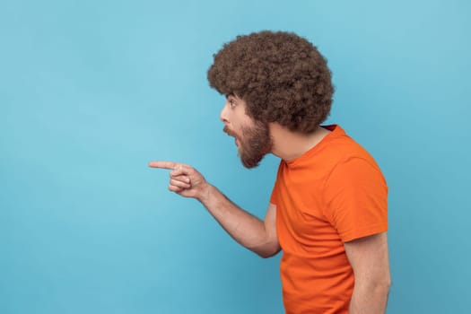 Side view of man with Afro hairstyle in orange T-shirt pointing aside, showing blank copy space for idea presentation, commercial text, looking ahead. Indoor studio shot isolated on blue background.
