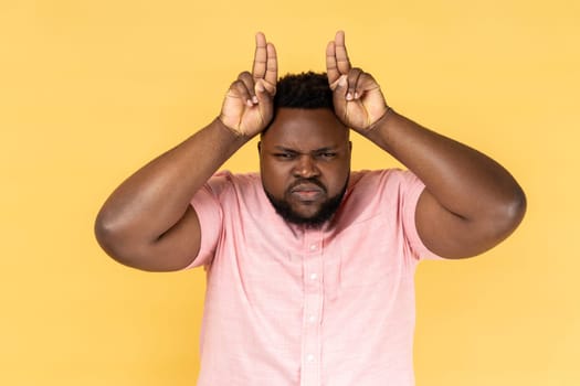 Portrait of man wearing pink shirt showing bull horn gesture, looking at camera with hostile angry expression, bully male threatening to attack. Indoor studio shot isolated on yellow background.
