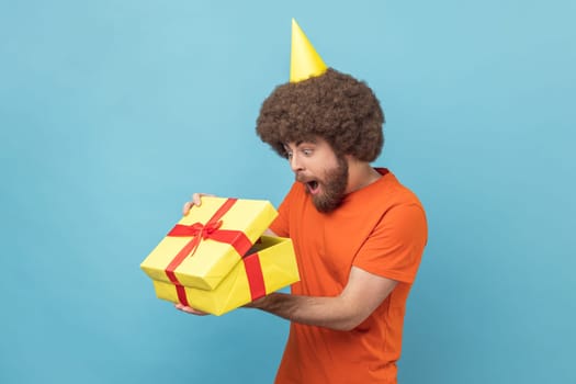 Portrait of surprised man with Afro hairstyle wearing orange T-shirt and party cone unpacked present box, looking inside with open mouth. Indoor studio shot isolated on blue background.