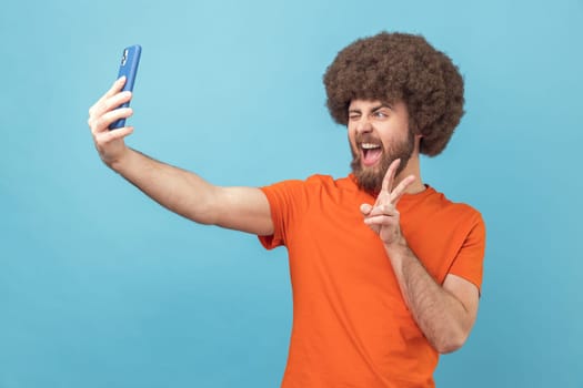 Portrait of man with Afro hairstyle wearing orange T-shirt showing v gesture, posing at phone camera, making selfie or having video call. Indoor studio shot isolated on blue background.