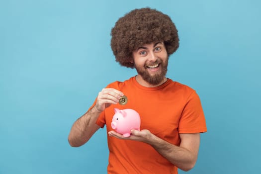 Portrait of man with Afro hairstyle wearing orange T-shirt investing in bitcoins, putting golden crypto coin into piggy bank, happy with money savings. Indoor studio shot isolated on blue background.