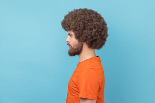 Side view of man with Afro hairstyle in T-shirt looking directly ahead with serious attentive face, listening attentively, unsmiling determined male. Indoor studio shot isolated on blue background