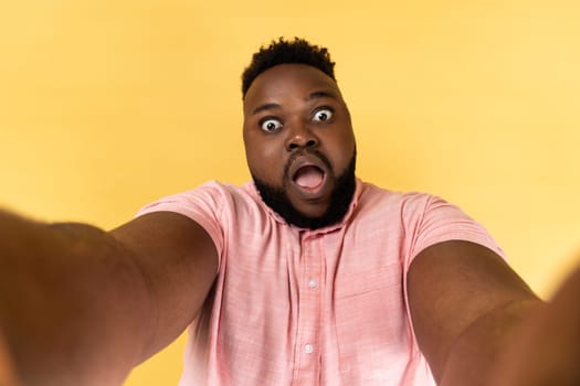 Portrait of shocked man wearing pink shirt making point of view photo, having surprised expression, POV, looking at camera with open mouth. Indoor studio shot isolated on yellow background.