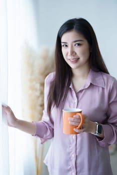 A portrait of a young Asian woman in pajamas smiling happily by a window with curtains in the morning sun while drinking coffee.