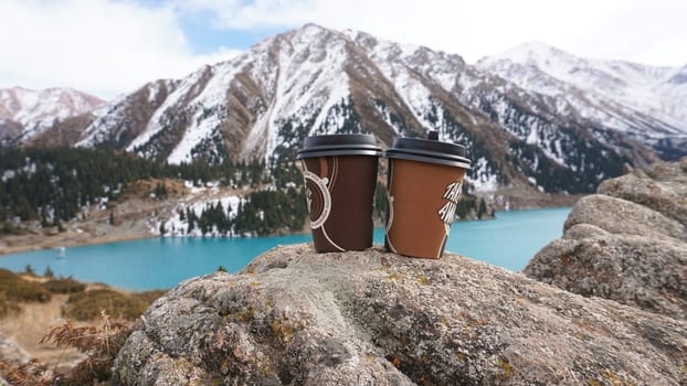 Paper cups with coffee on the background of a mountain lake. The color of the water is blue. A green forest grows on the hills. The high peaks are covered with snow. The cups are on a stone. Romance